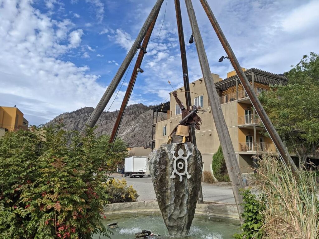 Water feature at Spirit Ridge, featuring Indigenous archer with tipi structure above. There is a four storey building in the background