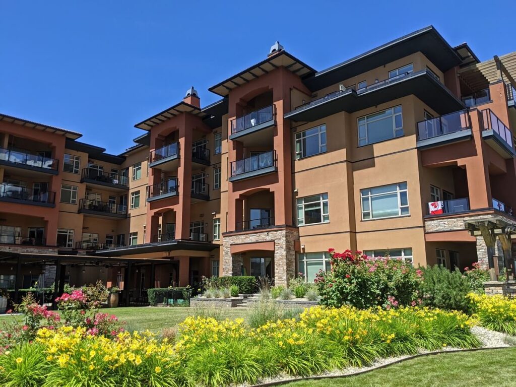 Exterior shot of Watermark Resort in Osoyoos - a tan coloured four storey building with large windows and balconies. There are colourful flowers in front of the building.