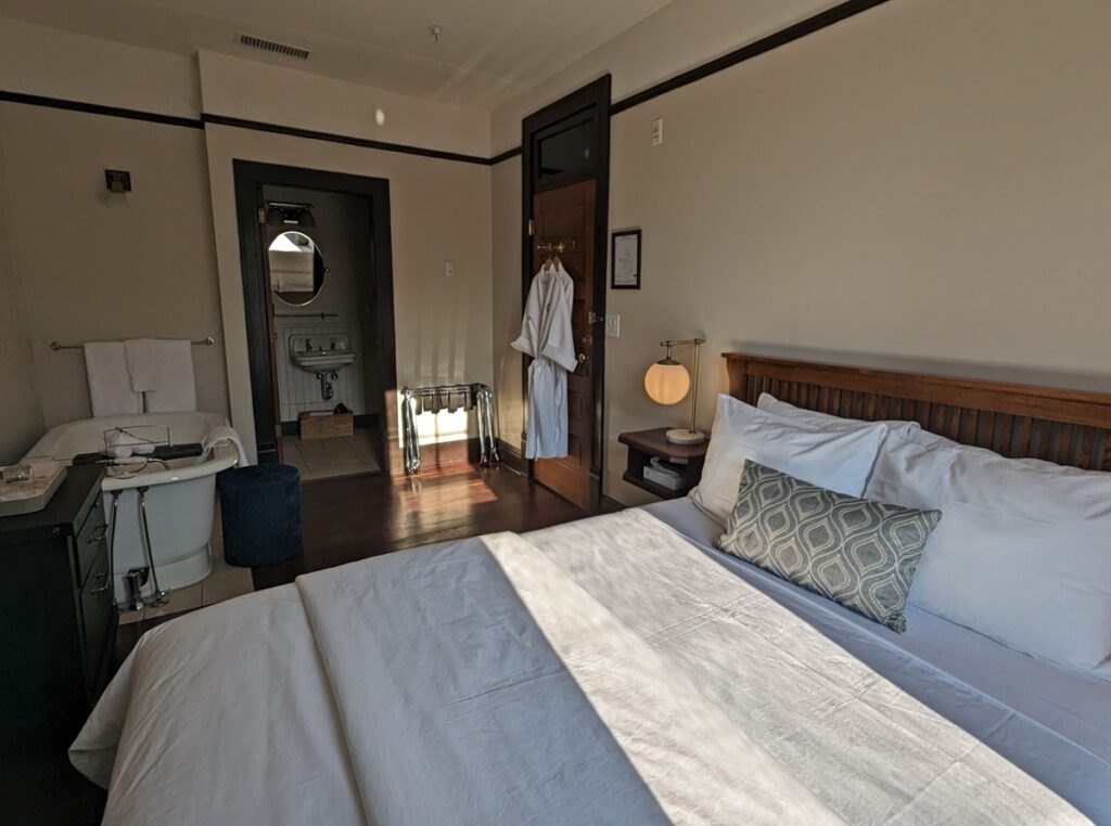 Bedroom view at the Naramata Inn, featuring large queen bed, rolltop bath on left and bathroom visible. There are robes on the back of the door