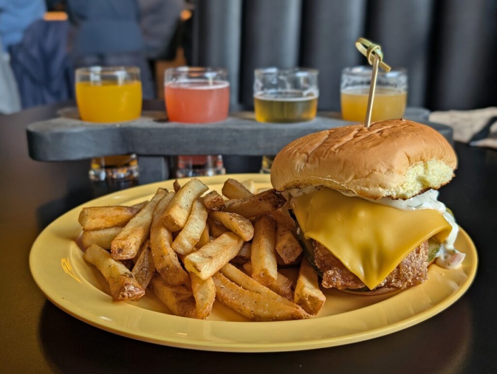 Close up of burger at Yellow Dog Brewing, with the patty, bun and cheese on right, fries on left. There is a flight of beers in the background