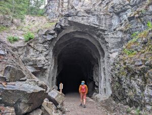 Back view of hiker in orange jacket with red backpack heading into tall southern entrance to Adra Tunnel