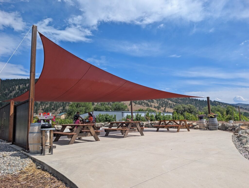 Side view of paved picnic patio at Three Sisters Winery with large red sail shade above picnic tables, surrounded by trees and vineyards