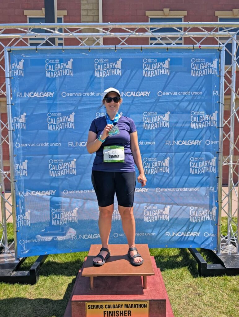 Gemma is standing in front of Calgary Marathon signage, wearing running gear and holding a marathon finisher medal