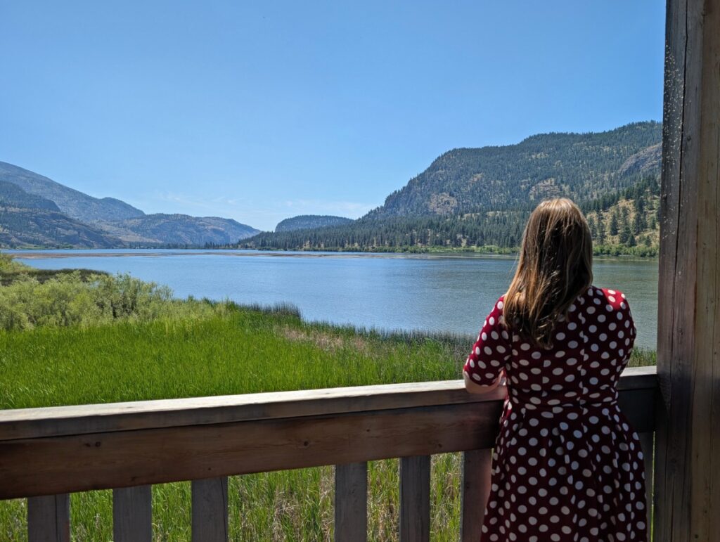 Back view of woman in red and white poka dot dress in Vaseux lake bird hide, looking out to calm lake surrounded by forested mountains