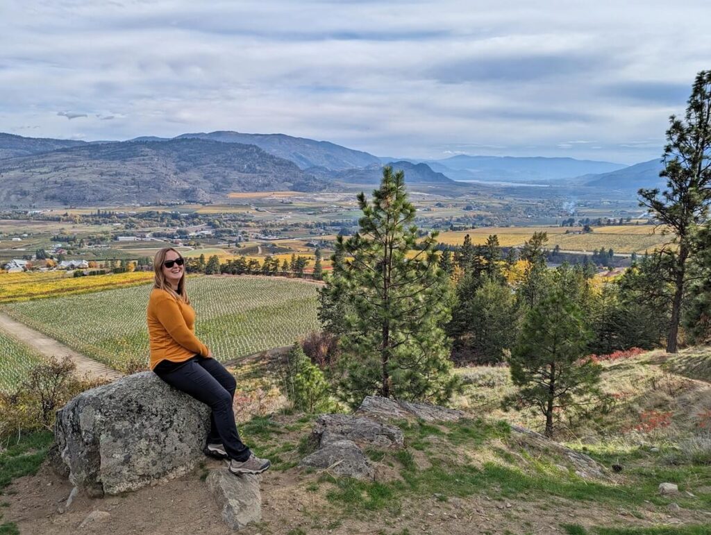 Gemma sits on a wall in front of views of vineyards and mountains on the Golden Mile Trail in Oliver