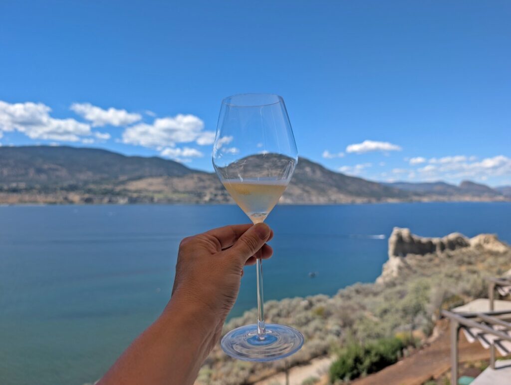 A hand holds up a glass of sparkling wine at Evolve Sparkling House in Penticton, with a view of deep blue Okanagan Lake in the background, surrounded by forested hills