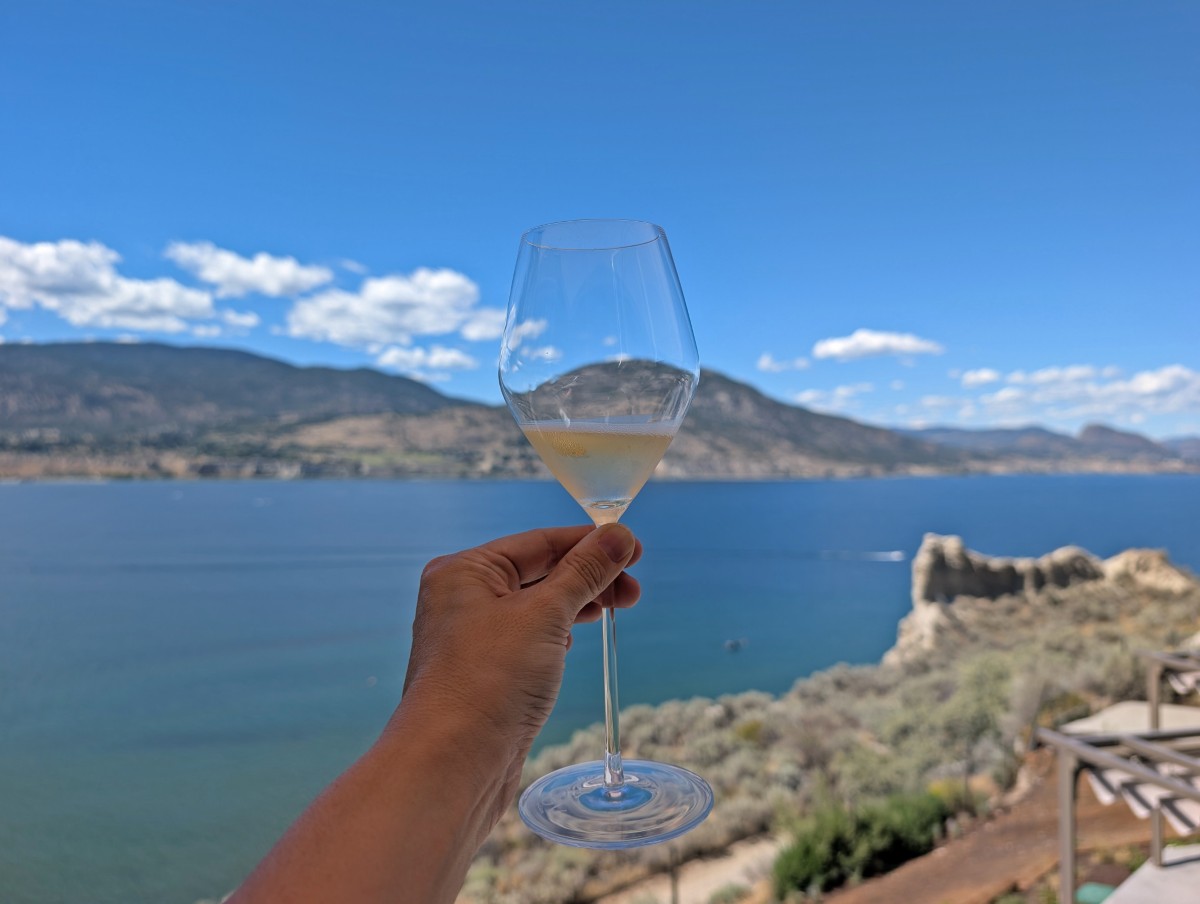 A hand holds up a glass of sparkling wine at Evolve Sparkling House in Penticton, with a view of deep blue Okanagan Lake in the background, surrounded by forested hills