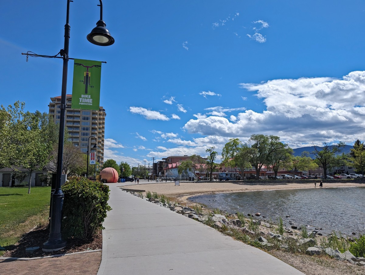 Street view of waterfront path along calm lake in Penticton, with sandy beach and Peach visible ahead